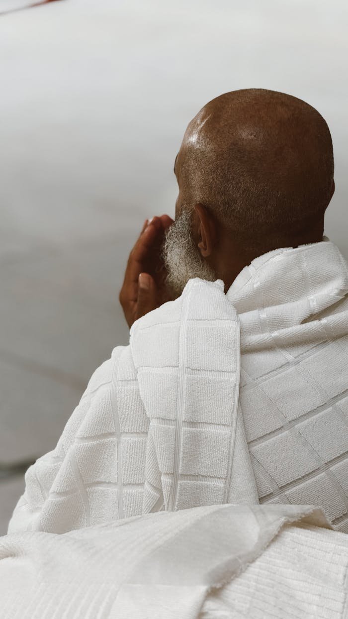An elderly man in a white robe prays with deep concentration, seen from behind.