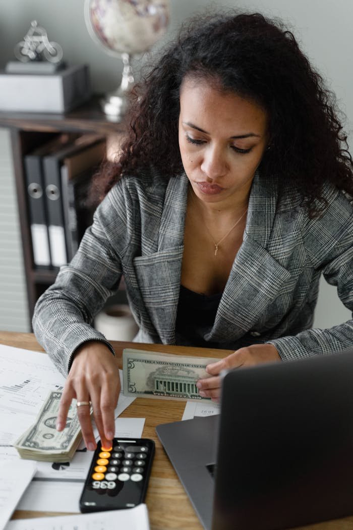 Focused businesswoman handling cash and calculations at her office desk, wearing a plaid blazer.