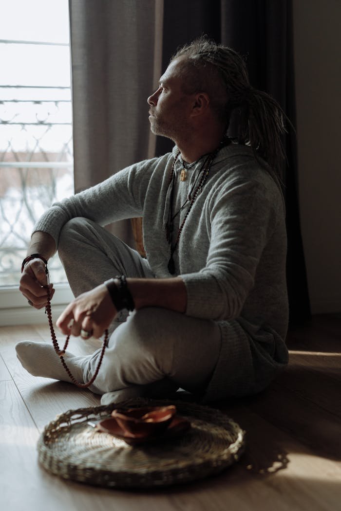 Man in natural light holding prayer beads, sitting indoors in deep thought.