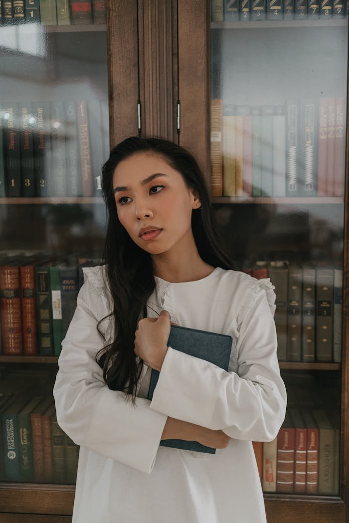 A woman in a white dress stands thoughtfully in front of bookshelves, holding a book.